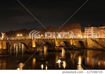 Illuminated Ponte Sant Angelo bridge over Tiber River in Rome at night with glowing reflections on water historic architecture and soft urban lights. High quality photo 127296728