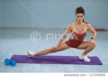 sporty girl is engaged in a left lunge stretch on a purple fitness mat, enhancing her flexibility and strength indoors She focuses on her form in a well lit gym environment 127299092