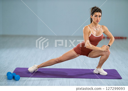 sporty girl executes left lunge stretch on purple yoga mat in gym She focuses on her form while surrounding weights lie on floor beside her 127299093