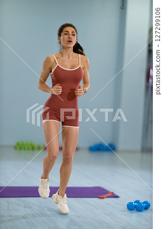 Focused on her fitness journey, a sporty girl is engaged in running in place She is inside a well lit gym with colorful weights and a fitness mat within reach, showcasing determination and energy 127299106
