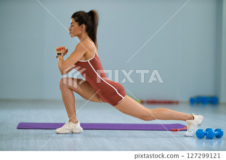 girl performs a stretching exercise by lunging to the right in a bright fitness studio. She focuses on her form while weights are visible in the background. Exercise mats line the floor. 127299121