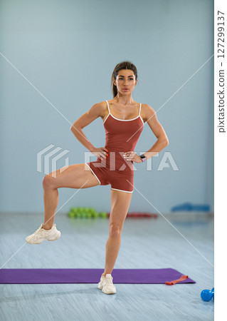 girl athlete is engaged in a standing side knee raise demonstrating her fitness skills in a well lit exercise studio. The setup includes workout equipment and a purple mat. 127299137