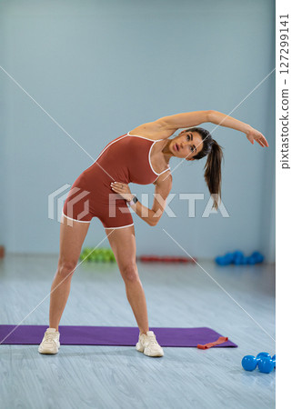girl athlete is bending her arm to the right and left while stretching in a gym setting. She demonstrates both flexibility and focus during her workout routine surrounded by exercise equipment. girl athlete is bending her arm to the right and left while stretching in a gym setting. She demonstrates both flexibility and focus during her workout routine surrounded by exercise equipment. 127299141