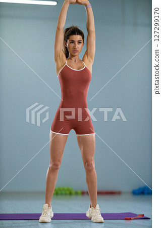 sporty girl is stretching her arms overhead as part of a warm up routine She is indoors, showcasing focus and determination in her fitness training, wearing a fitted outfit suitable for exercise 127299170