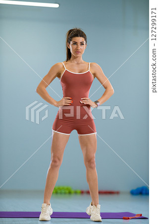 In a bright gym, a sporty girl is warming up with focused intensity She stands confidently with her hands on her hips, preparing for her workout routine to enhance agility and strength In a bright gym, a sporty girl is warming up with focused intensity She stands confidently with her hands on her hips, preparing for her workout routine to enhance agility and strength 127299171