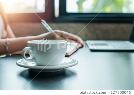 A serene morning scene featuring a cup of coffee, a woman writing in a notebook, and a laptop on a desk by the window. A serene morning scene featuring a cup of coffee, a woman writing in a notebook, and a laptop on a desk by the window. 127300046