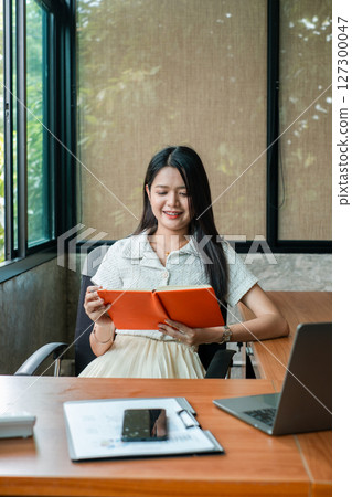 A young woman sits at a desk in a modern office, smiling while reading a tablet. Natural light filters through large windows. A young woman sits at a desk in a modern office, smiling while reading a tablet. Natural light filters through large windows. 127300047