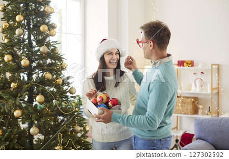 Happy funny in Santa hat and glasses decorating Christmas fir tree together at home 127302592