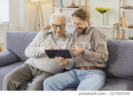 Senior man sitting on sofa at home with his adult son and using tablet together Senior man sitting on sofa at home with his adult son and using tablet together 127302593