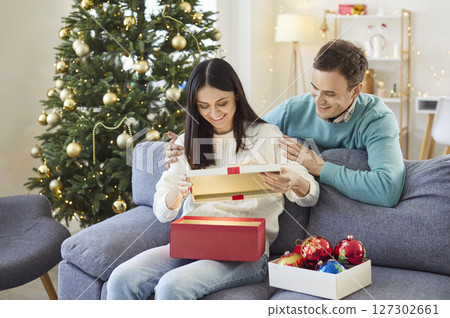 Happy young woman sitting on sofa at home with man opening red present box on Christmas holiday. 127302661