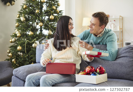 Happy young woman sitting on sofa at home with man opening red present box on Christmas holiday. 127302787
