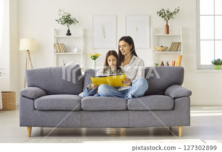 Young happy mother reading a book with her teenage daughter sitting on sofa at home. 127302899