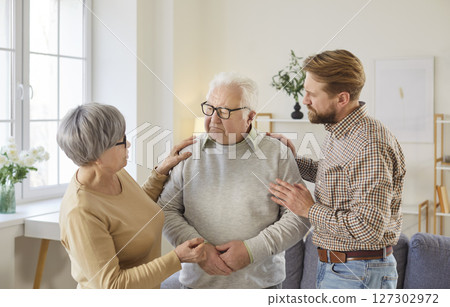 Senior woman and adult son supporting upset elderly man standing in living room at home together 127302972