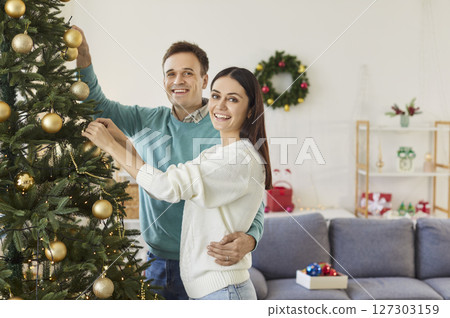 Loving happy couple at home decorating festive evergreen tree with balls for New Year, Merry Christmas 127303159