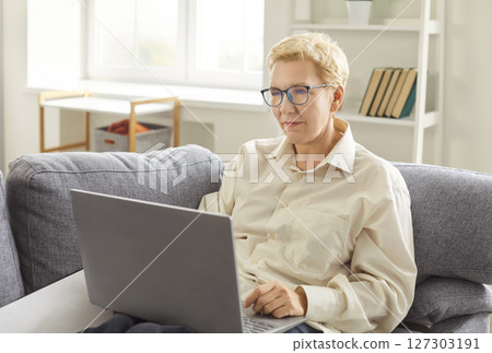 Portrait of confident senior woman using laptop sitting on couch in the living room at home. 127303191