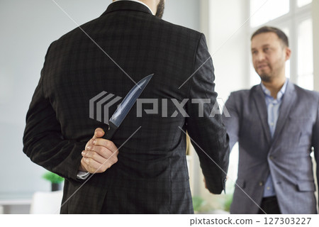 Man in suit hiding knife behind back while shaking hands symbolizing betrayal or backstabbing. 127303227