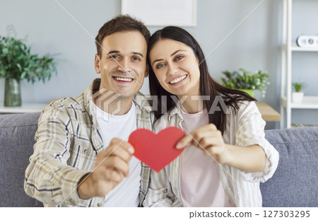 Young cheerful couple holding paper heart in hands sitting on sofa in living room at home. Young cheerful couple holding paper heart in hands sitting on sofa in living room at home. 127303295