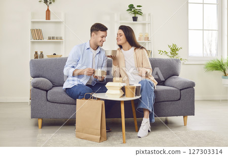 Couple eating takeaway food in boxes using delivery service sitting on sofa at home. Couple eating takeaway food in boxes using delivery service sitting on sofa at home. 127303314