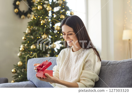 Young happy girl opening red present gift box sitting on sofa with decorated Christmas tree at home Young happy girl opening red present gift box sitting on sofa with decorated Christmas tree at home 127303320
