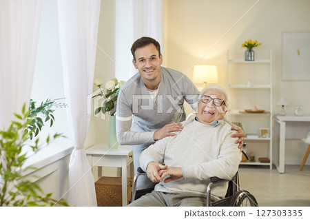 Young happy social worker standing with senior elderly disabled man with disability in wheelchair. Young happy social worker standing with senior elderly disabled man with disability in wheelchair. 127303335