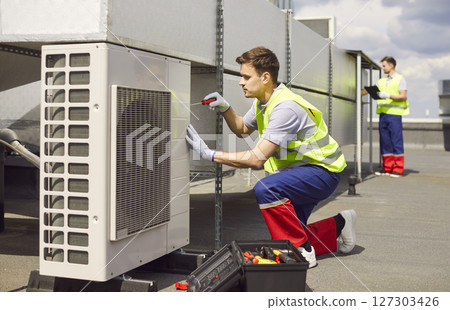 Technician installing and fixing air conditioner using screwdriver sitting on the roof of building. 127303426