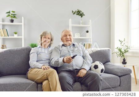 Senior couple watching tv holding TV remote and football ball supporting sport team together. 127303428