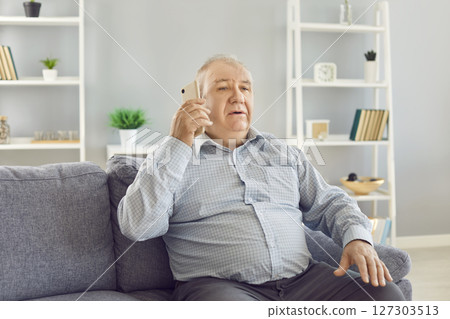 Serious Elderly Man Talking On Phone While Sitting On Sofa In Living Room Serious Elderly Man Talking On Phone While Sitting On Sofa In Living Room 127303513