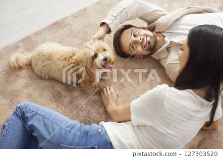 Cheerful couple lying on floor, playing with Maltipoo dog at home, enjoying joyful time together. Cheerful couple lying on floor, playing with Maltipoo dog at home, enjoying joyful time together. 127303520