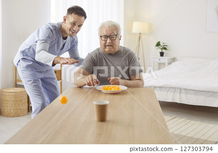 Senior man enjoying playful moment with cheerful caregiver, aiming ping pong balls into paper cup. Senior man enjoying playful moment with cheerful caregiver, aiming ping pong balls into paper cup. 127303794