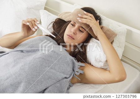 Sick young woman lying on bed in bedroom suffering from headache and measuring temperature 127303796