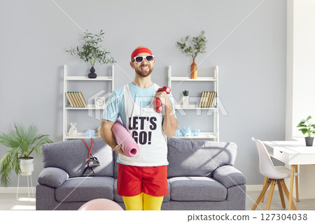 Funny man holding yoga mat and dumbbells and looking at camera standing in living room at home. Funny man holding yoga mat and dumbbells and looking at camera standing in living room at home. 127304038