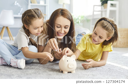 Family of mother and two kids putting money coin into piggy bank lying on floor at home together. Family of mother and two kids putting money coin into piggy bank lying on floor at home together. 127304174