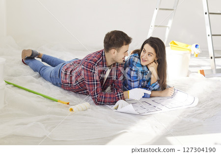 Couple lying on floor with blueprint and paint rollers discussing design project of new house. 127304190
