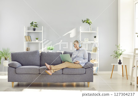 Senior woman using laptop and smiling while sitting on couch in at home in headphones. Senior woman using laptop and smiling while sitting on couch in at home in headphones. 127304237