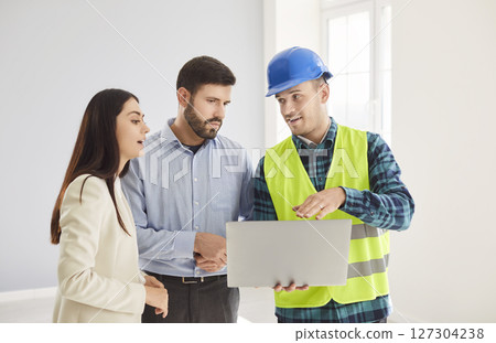 Construction worker in blue helmet showing apartment renovation plan on laptop to young couple. 127304238
