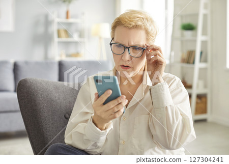 Senior shocked concerned cellphone female user staring at screen, elder woman at home 127304241