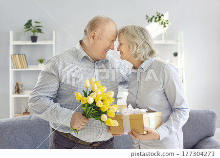 Happy senior couple standing at home with flowers bouquet and present gift box. Happy senior couple standing at home with flowers bouquet and present gift box. 127304271