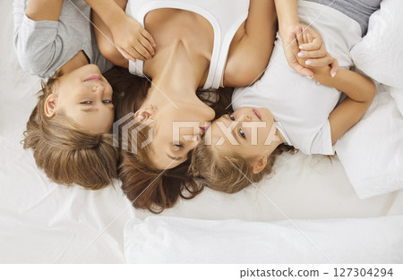 Top view of happy mother and her smiling children lying in bed, holding hands and sharing hugs. Top view of happy mother and her smiling children lying in bed, holding hands and sharing hugs. 127304294