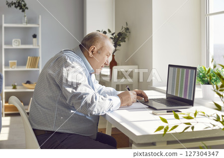 Old senior man accountant working using laptop with chart sitting at desk at home 127304347