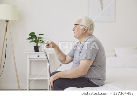 Portrait of a senior elderly man sitting on bed in bedroom at home with his crutch 127304492
