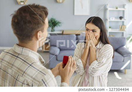 Young man with a red gift box proposing to his happy girlfriend standing in living room at home. 127304494