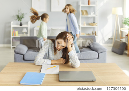 Frustrated mother sitting at desk, struggling to focus and work while noisy kids playing loudly. 127304500