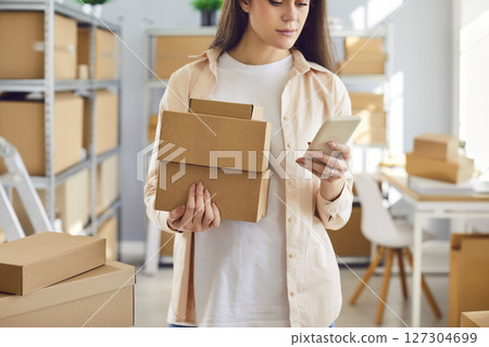 Young female employee holding cardboard boxes standing in warehouse with mobile phone. Young female employee holding cardboard boxes standing in warehouse with mobile phone. 127304699