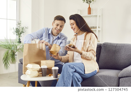 Young happy couple talking and eating take-away food in living room at home having lunch together. 127304745