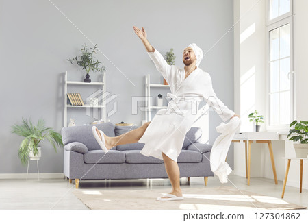 Happy funny young man in white bathrobe holding towel and having fun enjoying spa day at home. 127304862
