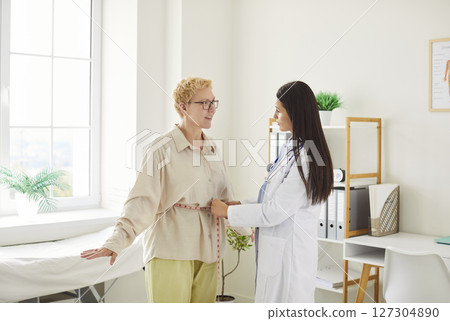 Young woman doctor measuring waist of female patient with tape measure during examination in clinic. Young woman doctor measuring waist of female patient with tape measure during examination in clinic. 127304890
