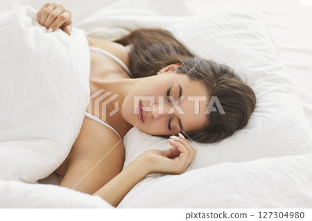 Young woman lying in cozy bed with closed eyes, resting peacefully under blanket in bright bedroom. Young woman lying in cozy bed with closed eyes, resting peacefully under blanket in bright bedroom. 127304980