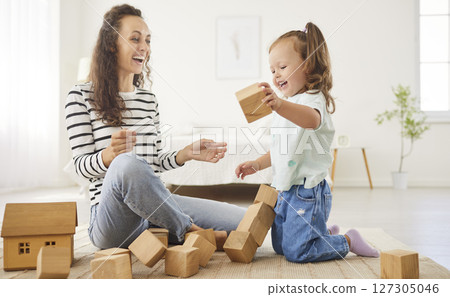Joyful mother and daughter laughing together after knocking down wooden block tower during playtime. 127305046