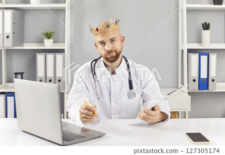 Funny young male doctor in white medical uniform sitting on workplace in clinic wearing crown. Funny young male doctor in white medical uniform sitting on workplace in clinic wearing crown. 127305174