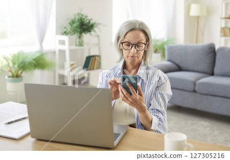 Senior woman working at the desk with laptop holding mobile phone in hands. 127305216